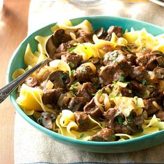 a blue bowl filled with pasta and meat on top of a wooden table next to a fork