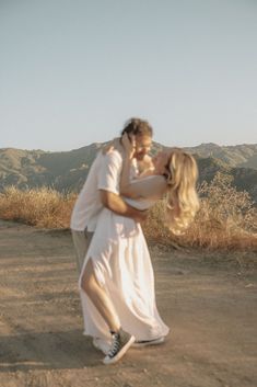 a man and woman kissing in the middle of a dirt road with mountains in the background