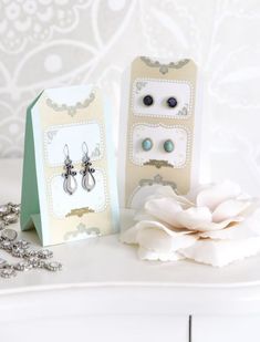 two small boxes with earrings on them sitting next to a white flower and necklaces