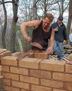 a man is building a brick wall in the woods while another man watches from behind