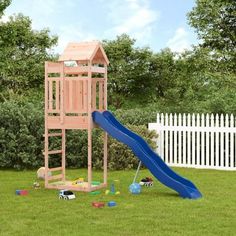 a wooden playset with a blue slide in the grass next to a white picket fence