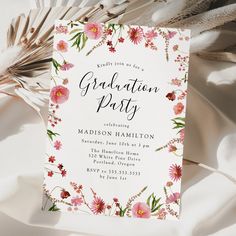 a white and pink floral graduation party card on top of a table with feathers in the background