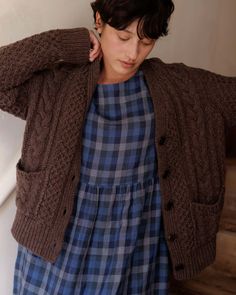 a woman in a blue dress and brown cardigan is leaning against a white wall