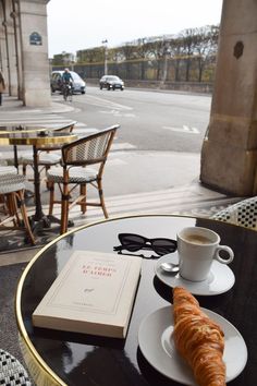 a table with a book, coffee cup and croissant sitting on top of it