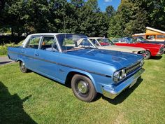 an old blue car is parked in the grass near other older cars and trucks behind it