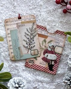 two tags with christmas images on them sitting next to pine cones and evergreens in the snow