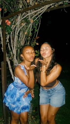 two young women standing next to each other in front of a tree and grass area