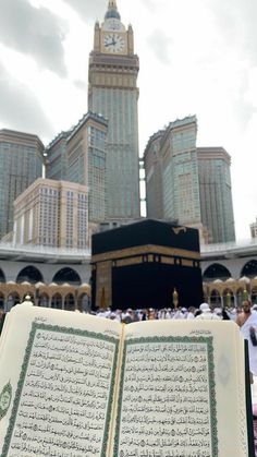 an open book with arabic writing in front of a clock tower and other tall buildings