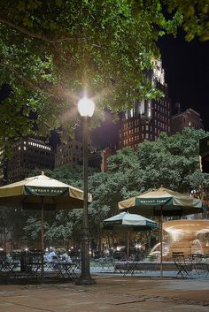 tables and umbrellas are lit up at night in a city park with tall buildings