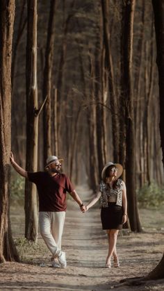 a man and woman holding hands walking down a dirt road in the woods with trees