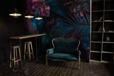 a chair and table in front of a wall with tropical leaves on it, next to a bookcase