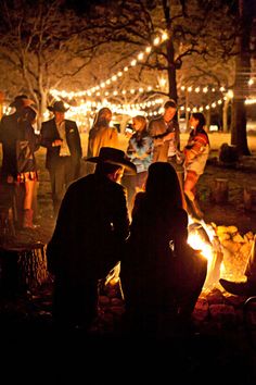 people sitting around a campfire at night with lights strung over the trees and onlookers