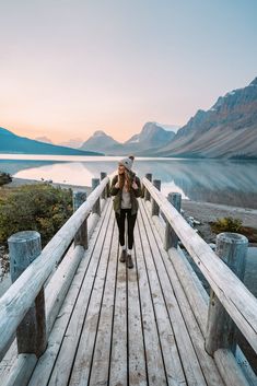 a woman walking across a wooden bridge over a body of water with mountains in the background