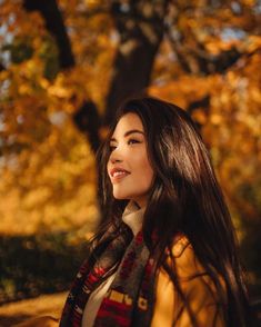 a woman with long brown hair wearing a plaid scarf and smiling at the camera while standing in front of a tree