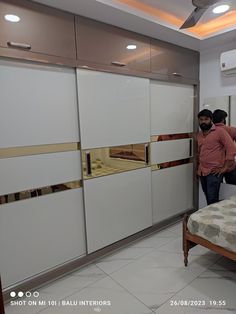 two men are standing in the corner of a room with white cupboards and a bed