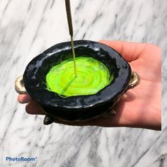 a hand holding a black bowl with green liquid in it and a stick sticking out of the top