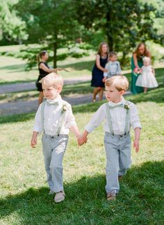 two young boys are walking in the grass holding hands and wearing suspenders with flowers on them