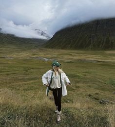 a woman walking across a lush green field under a cloudy sky with mountains in the background