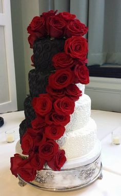 a three tiered wedding cake with red and black roses on the top, sitting on a table in front of a window