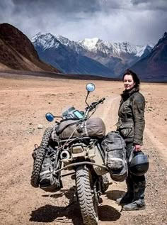 a woman standing next to a motorcycle on a dirt road with mountains in the background