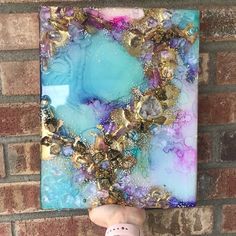 a person holding up a piece of art with gold and blue paint on it's face