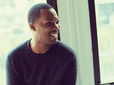 a man sitting in front of a window smiling at someone with his hand on his hip