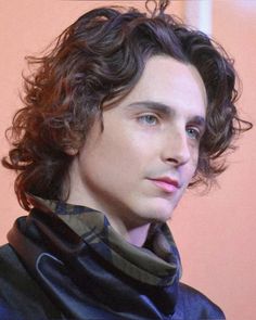 a man with curly hair wearing a blue shirt and scarf looking at the camera while standing in front of a pink wall