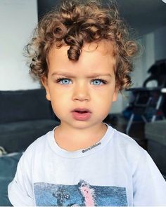 a little boy with curly hair wearing a t - shirt and looking at the camera