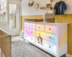 a white dresser with rainbow painted drawers and baskets on it's top, next to a yellow wall