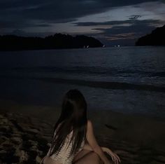 a woman sitting on the beach at night with her legs crossed and looking out into the water