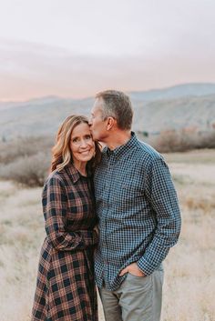 a man and woman are standing together in the grass with mountains in the back ground