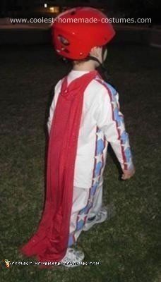 a young boy wearing a red helmet and cape standing in the grass with his hands on his hips