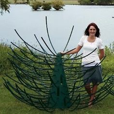 a woman standing next to a large metal peacock sculpture in front of a body of water