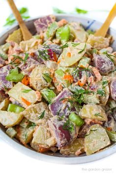 a bowl full of potato salad with two wooden spoons in the side and green herbs on top