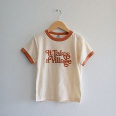 It Takes a Village, retro-inspired ringer tee inspired by the power of community, designed for all of us. Available for adults and kids. NOTE: - Runs True To Size - Not preshrunk so they do have about 3% shrinkage if dried. - Dyed using non-toxic dyes. - Screen printed using eco-friendly water-based inks. Each tee is made in Bangladesh by makers whose mission is to protect the environment and consciously strive to minimize the use of natural resources in the manufacturing industry. The factory i Ringer Tee Design, Retro Ringer Tee, Holiday Graphic Tee, 70s Ringer Tee, Retro Tshirt Design, 70s Tshirt, Merch Inspiration, Vintage Tshirt Design, Fun Tshirt