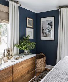 a bedroom with dark blue walls and white drapes on the windowsill, a large plant in a wicker basket