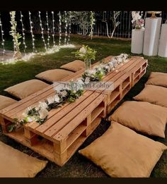 a wooden table with lots of pillows and flowers on it in the middle of some grass