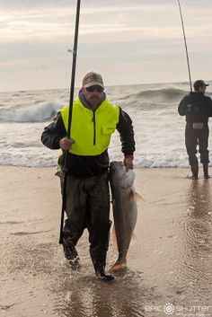 a man holding a fish while standing on top of a beach next to the ocean