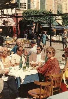 an old photo of people sitting around a table eating food and drinking wine in the street