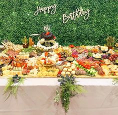 a table topped with lots of food next to a wall covered in greenery and happy birthday sign