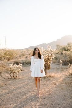 a woman in a white dress walking through the desert with cacti behind her