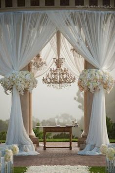 an open window with white drapes and flowers on the windowsill, in front of a chandelier