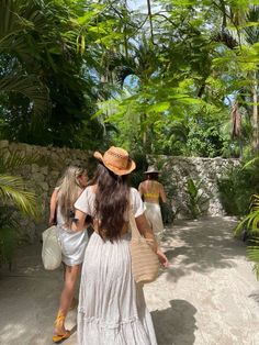 two women in white dresses and hats walking down a path with palm trees on either side