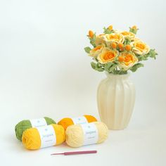 three balls of yarn next to a white vase with yellow flowers and crochet needles