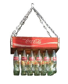 an old coca - cola sign hanging from a chain with six empty coke bottles in it