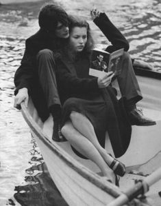 two people sitting in a boat on the water, one reading a book while the other looks at something