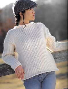 a woman wearing a hat and sweater leaning on a fence with her hands in her pockets