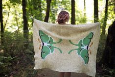 a woman standing in the woods holding up a blanket that has two green flowers on it