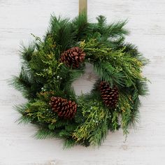 a green wreath with pine cones hanging on a wooden door hanger against a white painted wall