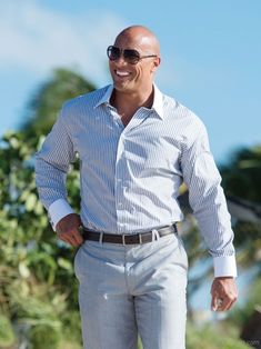 a bald man wearing sunglasses and a white shirt smiles at the camera while standing in front of some trees